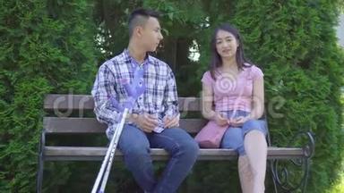 年轻积极的亚裔男子，拄着拐杖断腿，与坐在长凳上的年轻亚裔女子交流